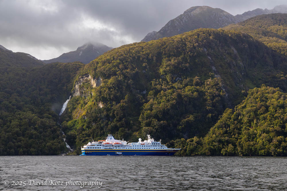 a cruise ship in the water, with a forested hillside behind, and a waterfall cascading down that hillside, and mountain peaks beyond.