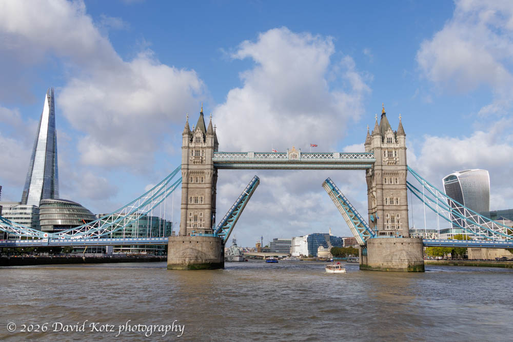 A view from the river of Tower Bridge, open for boat traffic, with the Shard at left and another iconic skyscraper at right.