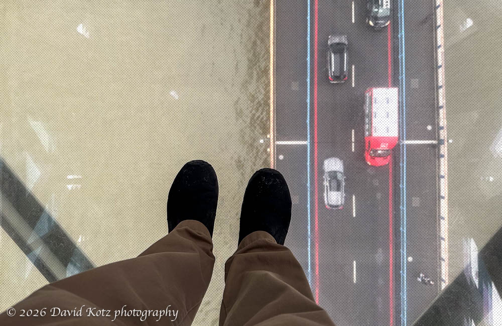 looking down at my feet, on a glass floor in the high/pedestrian walkway connecting the towers on Tower Bridge.  we can see traffic on the road below, and the river below that.